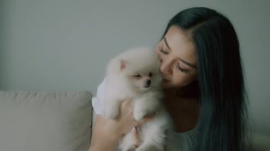 Genç Taylandlı bir kadın kucağında küçük şirin beyaz bir köpek, oturma odasında koltuğunda oturuyor, uzun saçlı genç bir kadın küçük tüylü bir köpek tutuyor ve gülümsüyor. İnsanlar ve hayvanlar arkadaşlık kavramı. Yüksek