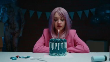 Üzgün, pembe saçlı, makyajlı ve pembe ceketli bir kız. Masada oturmuş, mavi doğum günü pastasıyla ağlıyor. Karanlık bir odada doğum günüyle mum yakıyor.