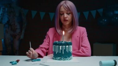 Üzücü pembe saçlı, makyajlı, pembe ceketli doğum günü pastası olan kız masada oturuyor ve doğum günü süslemeleri olan karanlık bir odada mum yakıyor. Yüksek kalite 4k görüntü