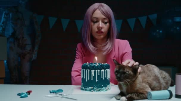 青い誕生日のケーキとテーブルに座って 誕生日の装飾で猫を暗い部屋に置くピンクのブレザーでメイクした悲しいピンクの髪の誕生日少女 高品質の4K映像 — ストック動画