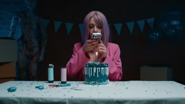 悲伤的粉红头发的生日女孩穿着粉红夹克坐在桌旁 用一支蜡烛在一间装饰着生日装饰品的黑暗房间里为蓝色的生日蛋糕拍照 — 图库视频影像