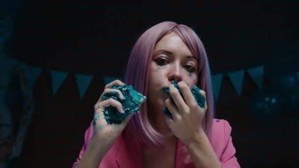 悲伤的粉红头发的生日女孩穿着粉红夹克坐在桌旁 拿着一大块蓝色的生日蛋糕 双手捧着生日装饰品在一间黑暗的房间里吃着 — 图库视频影像