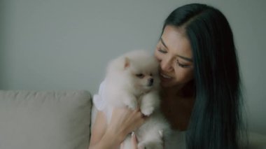 Genç Taylandlı kadın portresi, oturma odasındaki kanepede oturan, küçük tüylü bir köpek tutan ve gülümseyen, küçük, siyah, uzun saçlı bir kadın. İnsanlar ve hayvanlar dostluk