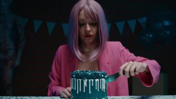 悲伤的粉红头发的生日女孩穿着粉红夹克坐在桌旁 用小刀切一个蓝色的生日蛋糕 用生日装饰在黑暗的房间里舔她的手指 — 图库视频影像