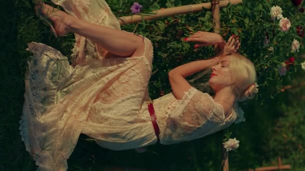 白い美しいヴィンテージレースの夏のマキシードレスの若いブロンドの女性の垂直ビデオ 晴れた日にはバラと花畑に座っています 黄金の時間に屋外でポジティブなブロンドの女性 — ストック動画