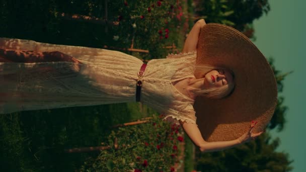 一个年轻的金发女子戴着宽边帽 身穿白色老式花边夏装的垂直视频 阳光灿烂的一天 站在玫瑰花园里 金光闪闪的金光闪闪的金发女人们肖像画 — 图库视频影像