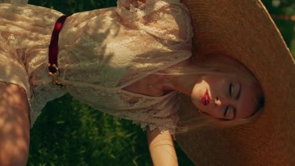 一个金发碧眼的年轻女子戴着宽边帽 穿着蕾丝夏装的垂直录像 在一个阳光灿烂的日子坐在玫瑰园里 金光闪闪的金光闪闪的金发女人们肖像导向 — 图库视频影像