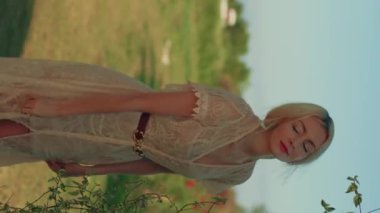 Beyaz, klasik bir yaz elbisesi içinde kırmızı rujlu, güzel, gülümseyen bir kadın. Güneşli bir günde doğada poz vermek. Açık havada sarışın bir kadın var. Portre oryantasyonu, dikey video. Yüksek kalite 4K