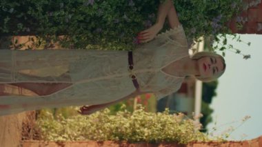 Beyaz, klasik bir yaz elbisesi içinde kırmızı rujlu, gülümseyen genç bir kadının dikey videosu. Güneşli bir günde doğada poz vermek. Altın saat içinde açık havada sarışın bir kadın. Portre