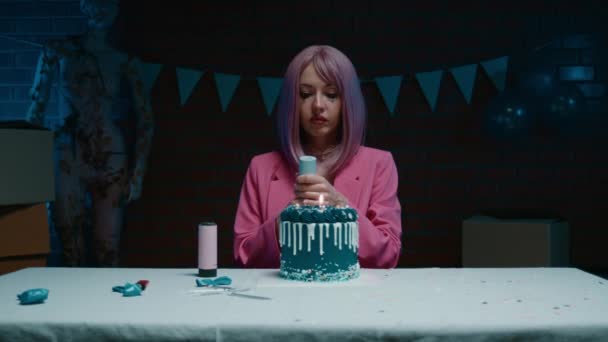 悲伤的粉红头发的生日女孩穿着粉红夹克坐在桌旁 手里拿着蓝色的生日蛋糕 在一间昏暗的房间里 放了一支彩蛋炮 吹灭了生日蜡烛 — 图库视频影像