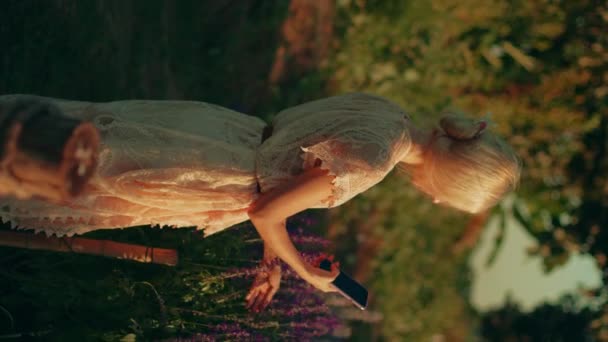 一个金发碧眼的年轻女子穿着白色蕾丝裙 带着智能手机在金光闪闪的紫罗兰花田里拍照 夏天的概念 大自然 野生野花 — 图库视频影像