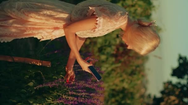 一个金发碧眼的年轻女子穿着白色蕾丝裙 带着智能手机在金光闪闪的紫罗兰花田里拍照 夏天的概念 大自然 野生野花 — 图库视频影像