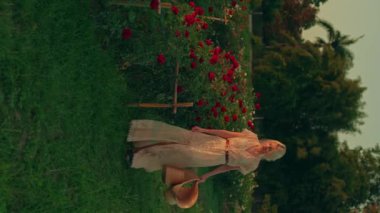 Beyaz dantel elbiseli ve geniş kenarlı hasır şapkalı genç sarışın bir kadın altın saatinde güzel kırmızı gül bahçesinde yürüyor. Yaz konsepti, doğa. Portre oryantasyonu, dikey video. Yüksek