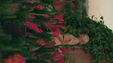 Beyaz boho elbiseli genç sarışın bir kadın kırmızı ve pembe çiçeklerle güzel renkli tarlada yürüyor. Yaz konsepti, doğa, kırsal alan. Portre oryantasyonu, dikey video. Yüksek