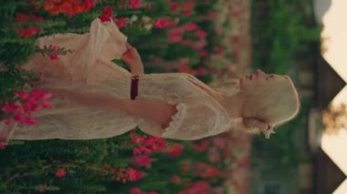 Beyaz dantel elbiseli genç sarışın bir kadın kırmızı ve pembe çiçeklerle güzel renkli tarlada yürüyor. Yaz konsepti, doğa, kırsal alan. Portre oryantasyonu, dikey video. Yüksek