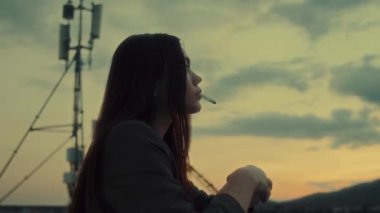 Çatıda sigara içen esmer bir kadının profil portresi. Gün batımında gökyüzü manzaralı. Özgürlük, gençlik, şehir manzarası 4K görüntüleri. Yüksek kalite 4k görüntü