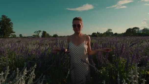 身穿白色夏装 戴着眼镜的雌性在浓密的高草草丛中穿过草地走向摄像机 在阳光灿烂的一天 她的手慢慢地触摸着花朵的顶部 高质量的 — 图库视频影像