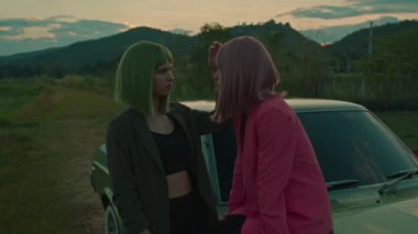 Yeşil saçlı kız, kız arkadaşının saçına dokunuyor. Pembe boyalı saçlı. Açık yeşil arabanın üzerinde oturuyor ve dağ manzaralı. Yavaş çekim