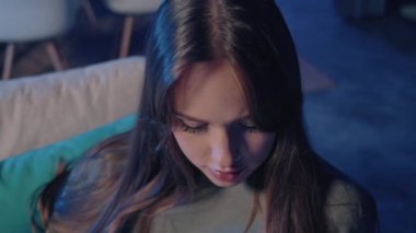 Uzun siyah saçlı bir kadın kanepede oturuyor, telefonuna bakarken gülümsüyor. Elektrikli mavi gözleri ve uzun kirpikleri kaldırdığı kaşlarını tamamlıyor ve sahneye bir neşe katıyor.