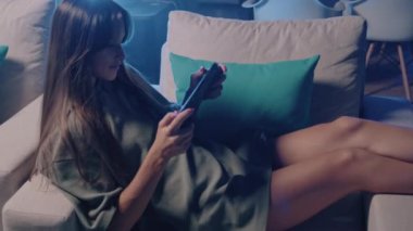 Bir kadın kanepede uzanmış parmak uçlarını kullanarak tablet bilgisayarla etkileşime geçiyor. Elektrikli mavi tırnak cilası ekrandaki yazı tipiyle çelişiyor. Bu da sakin ve eğlenceli bir atmosfer yaratıyor.