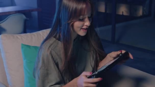 女性は 層状の髪と電気ブルーの服を着て 暗闇の中でソファに座っている間 彼女の太ももはやクッションの上に休息しながら 彼女の携帯電話でビデオゲームをプレイして楽しんでいます — ストック動画