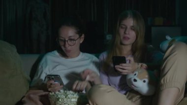 Pijamalarıyla oturup patlamış mısır yiyen ve geceleri rahat bir kanepede film izleyen iki genç kadının portresi. Karanlık odada televizyon ışığı var. Yüz ifadesi. Yüksek kalite