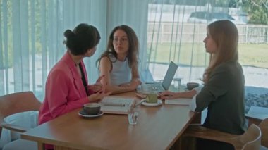 Modern uluslararası ofis. Kadınların iş buluşması. Üç kadın ortak çalışma alanında kahve ile fikirleri tartışıyor. İnsanlar masada oturuyor, beyin fırtınası yapıyor ve yaratıyorlar. Yüksek kalite 4k görüntü