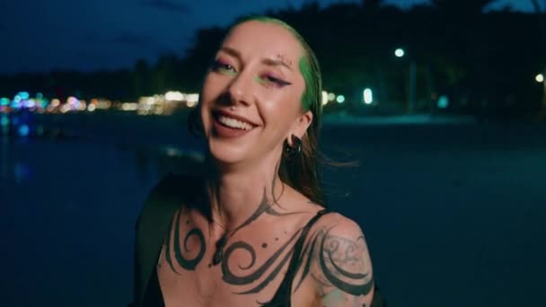 一个有着绿色头发和纹身的快乐女人 晚上站在蓝色的水边 嘴角挂着微笑 用她的大拇指做着一个悠闲的手势 享受着旅行的乐趣 — 图库视频影像