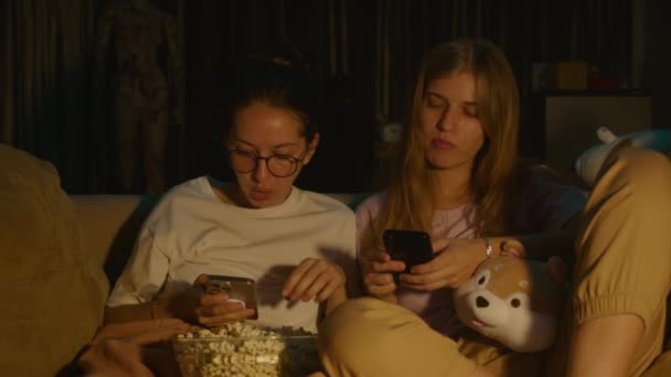 两个年轻女子穿着睡衣坐在家里舒适的沙发上看电视 坐在手机里吃爆米花的画像 黑暗房间里的电视灯 面部表情 高质量4K — 图库视频影像