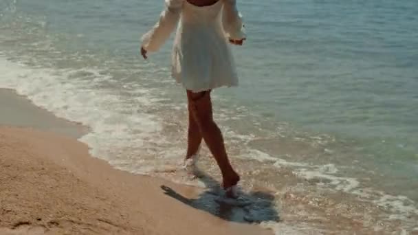 白いドレスの女性は 彼女が美しいビーチの風景を認めるように彼女の裸足の風波を感じ 液体の岸に沿って裸足を散歩しています — ストック動画