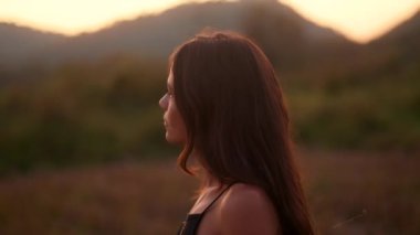 Siyah bir yaz elbisesi içindeki gülümseyen bir esmerin profil portresi. Gün batımında bir yaz gününde doğada poz veriyor. Yüksek kalite 4k görüntü