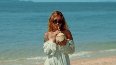 Beyaz elbiseli bir kadın, kumsalda gülümserken hindistan cevizinden gelen ferahlatıcı bir bardak suyun tadını çıkarıyor. Mutlu ve rahat görünüyor, güneş gözlüğü ve gözlük takıyor.