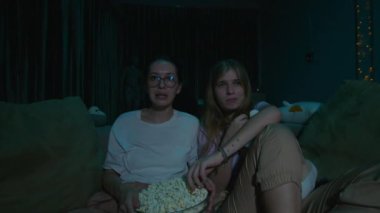Evdeki rahat bir kanepede oturup pijamalarıyla patlamış mısır yiyen iki genç kadın gece televizyon izlerken. Karanlık odada televizyon ışığı. Yüz ifadesi. Yüksek kalite 4k görüntü