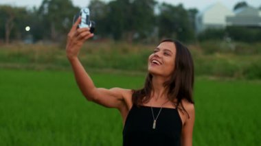 Genç ve çekici bir esmer, bir yaz akşamı pirinç tarlasında eski bir kamerayla selfie çekiyor. Tatil, boş zaman aktiviteleri, seyahat, hobi. Yüksek kalite 4k görüntü