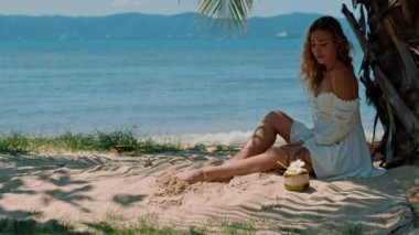 El değmemiş beyaz elbiseli bir kadın kumlu sahildeki bir palmiye ağacının altında mutlu bir şekilde oturur. Etrafı, doğanın ve güzel gökyüzünün keyfini çıkaran mutlu insanlarla çevrilidir.