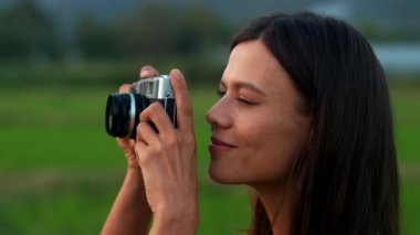 Bir yaz akşamı pirinç tarlasında gülümseyen ve eski bir kamerayla fotoğraf çeken çekici bir esmerin profil portresi. Tatil, boş zaman aktiviteleri, seyahat, hobi. Yüksek kalite 4k