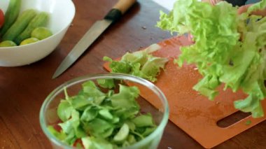 Bir insan, ahşap bir masaya yerleştirilmiş bir kesme tahtasının üzerinde marul doğruyor ve tarifin mutfağında bir yemek için malzeme hazırlıyor.