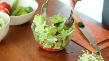 Bir insan, ahşap bir masada salata, marul, domates ve lahanayla besleyici bir salata hazırlarken görülüyor. Malzemeleri karıştırmak için kaşık ve çatal kullanıyor.