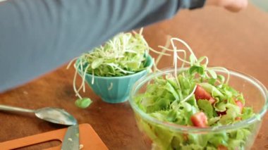 Bir insan, ahşap bir masada cam bir kasede taze sebze ve otları birleştirerek besleyici bir salata tabağı yaratıyor. Lezzet katmak için lahana ve erişte de ekliyorlar.