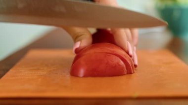 Genç bir kadın domatesleri bıçakla kesmek için tahta kesme tahtası kullanıyor. Parmakları ve başparmakları yemek malzemelerini dikkatlice tutar.
