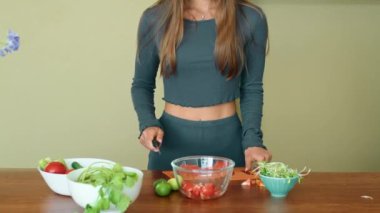 Bir kadın çeşitli sebzeler ve avokadoyu cam bir kaseye koyarak taze ve sağlıklı bir salata hazırlarken görülüyor. Taze meyvelerle dolu bir masanın önünde duruyor.