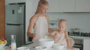 Bir ev hanımı ve bir bebek mutfakta mutlu bir şekilde yemek pişiriyorlar. Sarışın kadın güzellik kıyafeti giyerken, küçük kız şakacı hareketler yapıyor. Masa takımı ve mutfak eşyaları odayı dolduruyor.