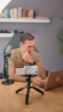 Bir adam evde çalışırken bulanık bir görüntüyle görülüyor. Çevrimiçi içerik oluşturmak için bir dizüstü bilgisayar ve mikrofon kullanıyor