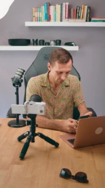 Çiçekli gömlekli bir adam bilgisayarında çalışıyor. Ofisteki masasında kamera ve mikrofon var..