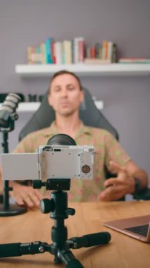 Bir adam, ahşap bir masada kamera, mikrofon ve laptop ile donatılmış bir stüdyoda podcast kaydediyor.