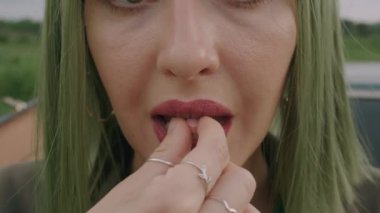 Yeşil peruklu genç bir kadın ağır çekimde eski bir arabanın bagajında oturmuş pembe hap yiyor.