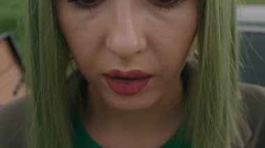 Yeşil peruklu genç bir kadın ağır çekimde eski bir arabanın bagajında oturmuş pembe hap yiyor.