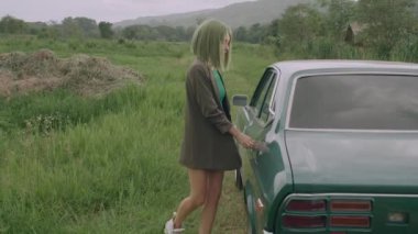 Yeşil peruklu şık bir kadın antika bir arabadan eski bir bavul çıkarır ve yavaş çekimde arabanın bagajına koyar.