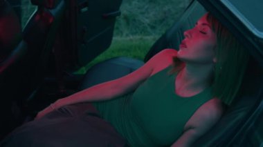 Yeşil elbiseli ve yeşil peruklu şık bir kadın, gözleri kapalı bir şekilde eski bir arabanın arka koltuğuna oturur.