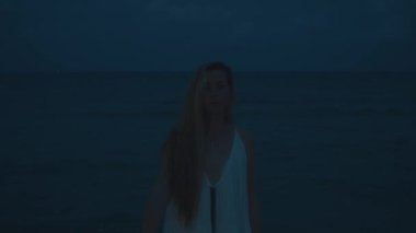 Geceleri deniz kenarında dalgalı saçları olan, elinde parlayan bir fener tutan bir kadın, huzurlu ve mistik bir atmosfer yaratır.